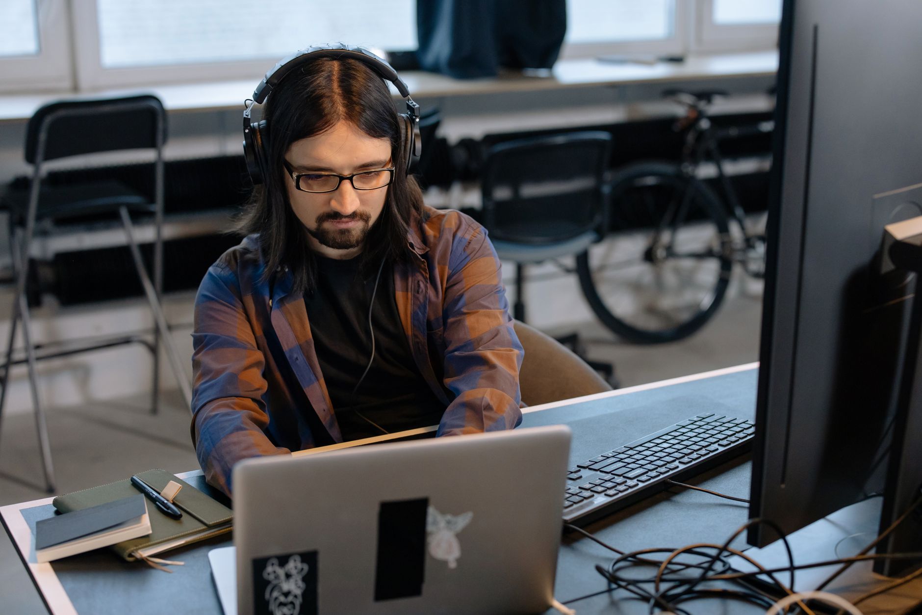 Homme avec casque audio et lunettes travaillant sur un ordinateur portable devant un grand écran dans un bureau lumineux.