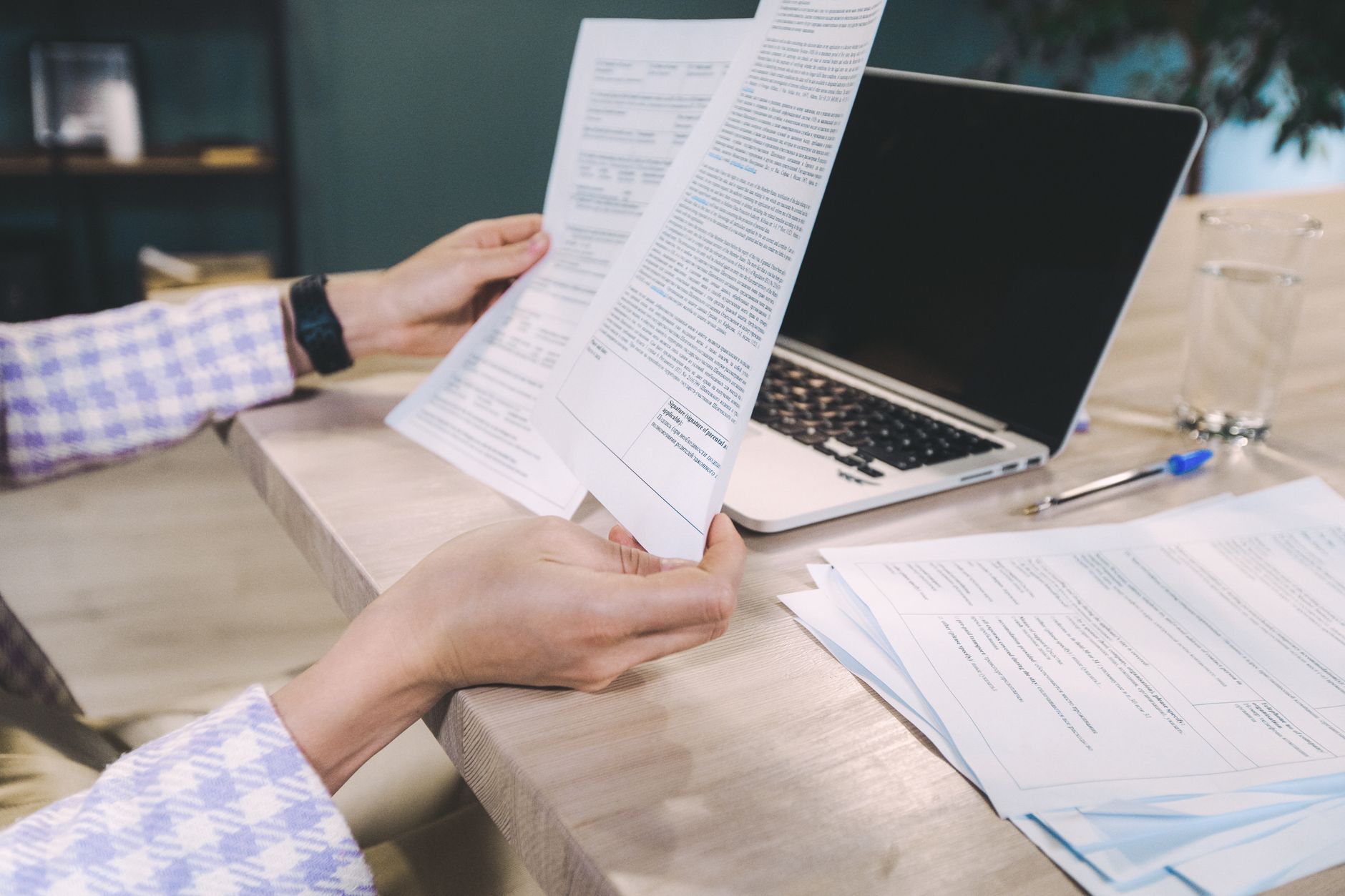 Mains tenant des documents imprimés devant un ordinateur portable sur une table de bureau.