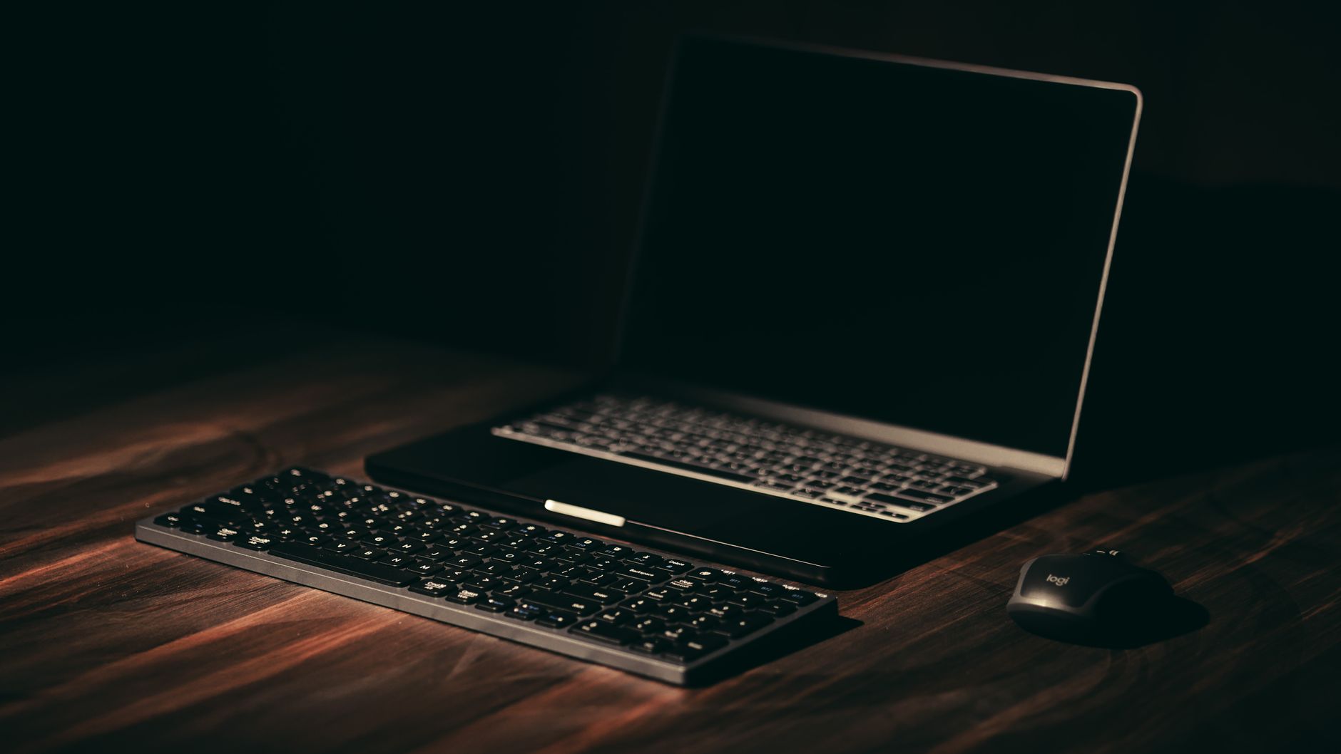 Ordinateur portable à l’écran éteint, clavier sans fil et souris sur un bureau en bois dans une pièce sombre.