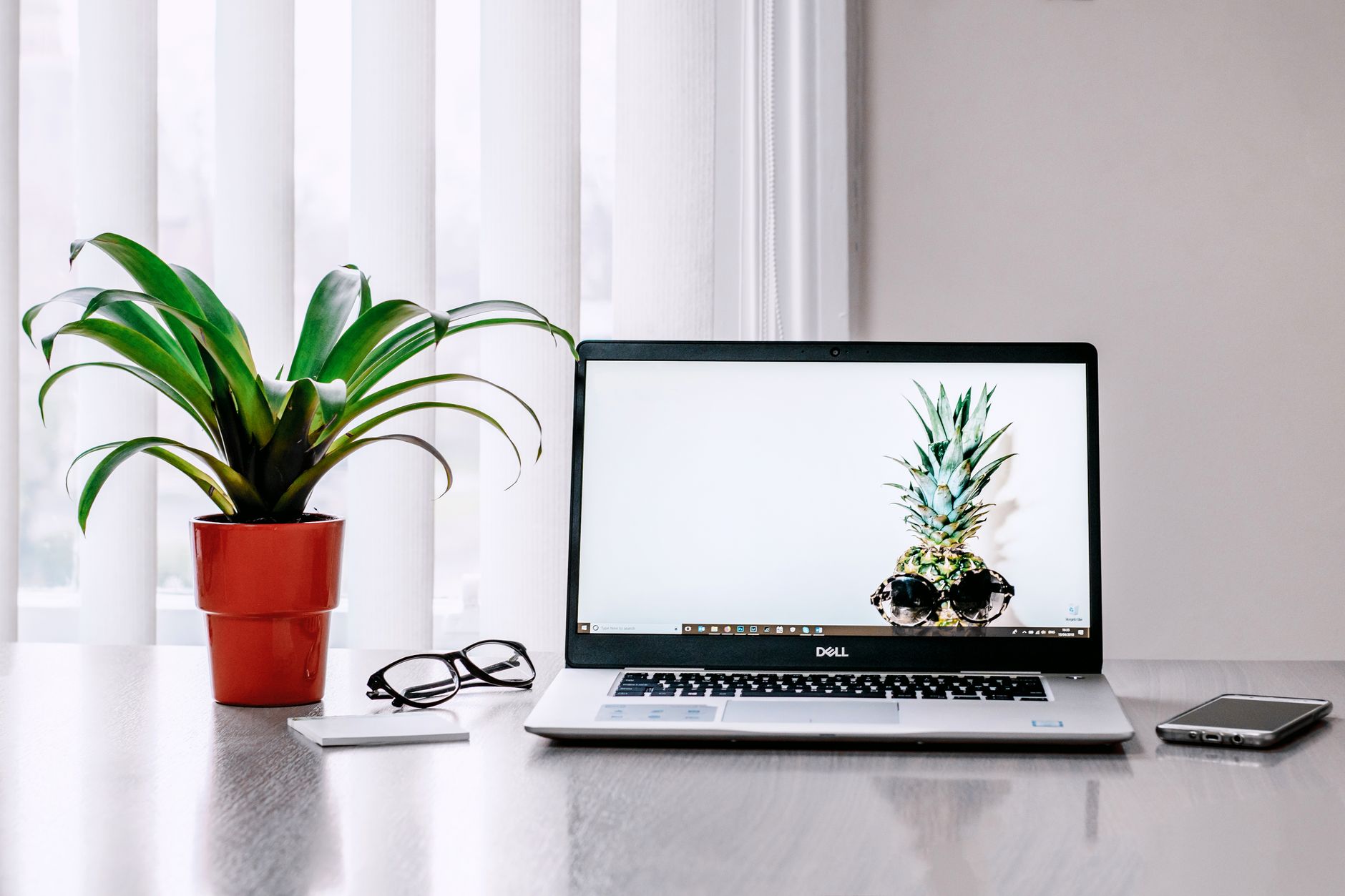 Ordinateur portable ouvert sur un bureau, à côté d’une plante en pot orange, de lunettes de vue et d’un smartphone, devant des stores clairs.