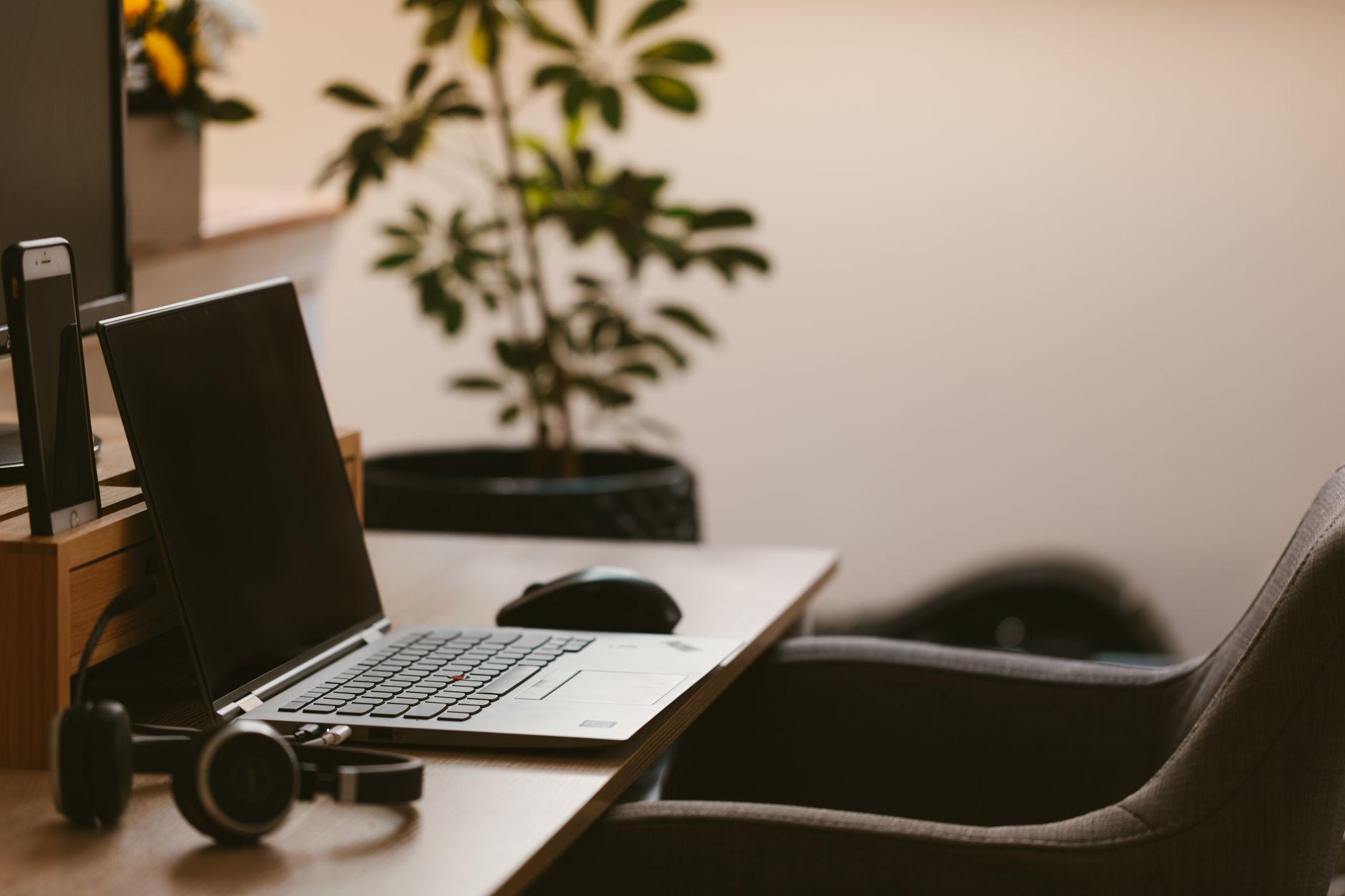 Ordinateur portable, souris et casque audio sur un bureau en bois, avec une plante verte et un fauteuil gris en arrière-plan